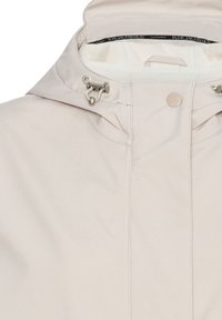 Světle béžová bunda s kapucí, kovovými zapínáními na šněrování a zapínáním na druky, uvnitř límce viditelná značka značky.