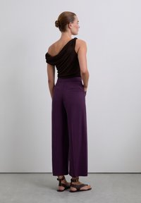 Brown draped top with one shoulder design, paired with high-waisted purple wide-leg pants. Brown studded sandals complete the outfit.