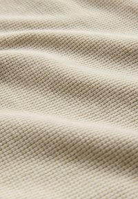 Beige tekstureret stof med et fint gitterlignende mønster, der viser bløde bølger og folder. Materialet ser blødt og vævet ud.