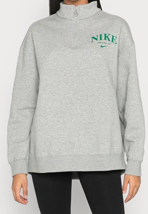 Persoon draagt een lichtgrijze Nike-pullover met een groen logo op de borst en een rits bij de nek, gecombineerd met zwarte legging.