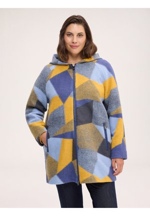 Woman wearing a hooded jacket with a blue, yellow, and gray geometric pattern, standing with hands in pockets against a plain background.