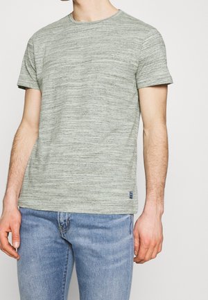 Mann trägt ein kurzärmeliges T-Shirt in Taubengrau und hellblaue Jeans und steht vor einem einfarbigen hellen Hintergrund.