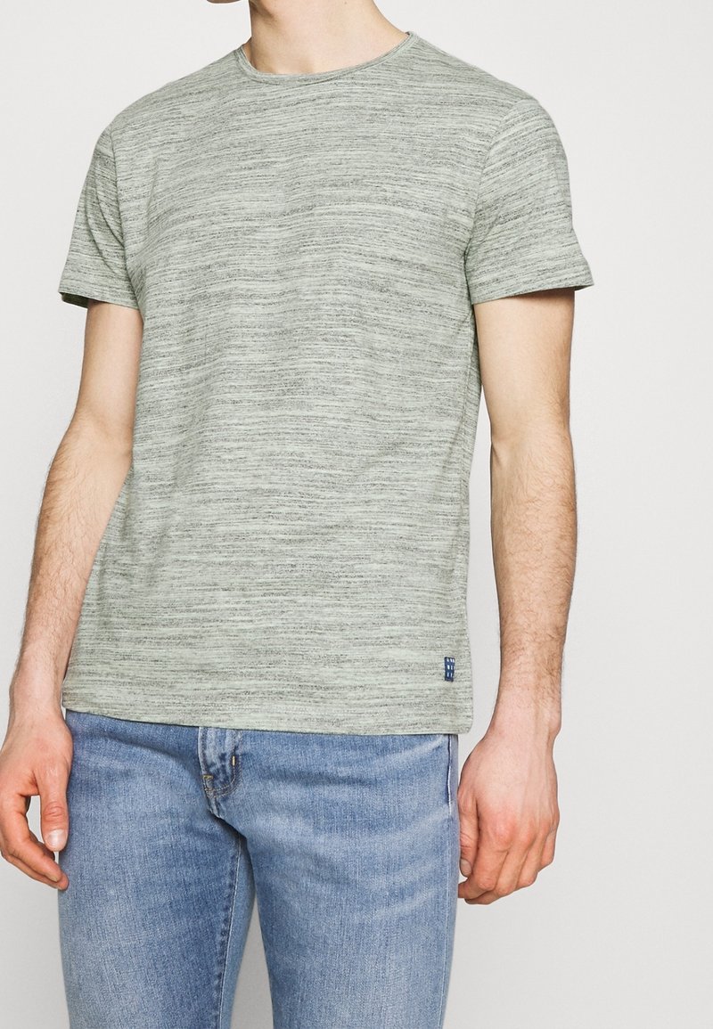 Homme portant un t-shirt gris chiné à manches courtes et un jean bleu clair, debout devant un fond clair uni.