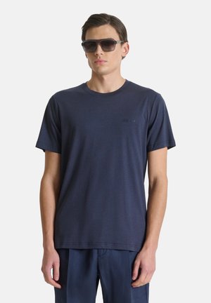 Giovane uomo che indossa una maglietta blu scuro e pantaloni coordinati, con capelli corti e grandi occhiali da sole neri, in piedi davanti a uno sfondo semplice.