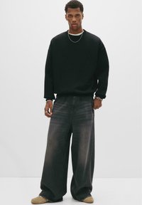 Sort crewneck sweatshirt, oversized pasform, stylet med brede grå jeans. Accessories inkluderer en kæd necklace og tynde armbånd.