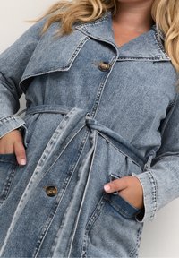 Detailní záběr osoby v džínové bundě s páskem a knoflíky, s rukama v předních kapsách a viditelnými vlnitými blond vlasy.