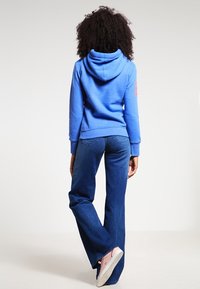 Felpa blu con cappuccio regolabile e dettaglio del logo sulla manica, abbinata a jeans a zampa d'elefante blu scuro e scarpe da ginnastica rosa. Tessuto dalla superficie liscia.