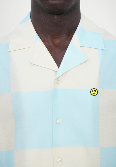 Camisa de manga corta con cuello en un patrón de cuadros en azul claro y beige, que cuenta con un pin de cara sonriente amarilla y botones transparentes.