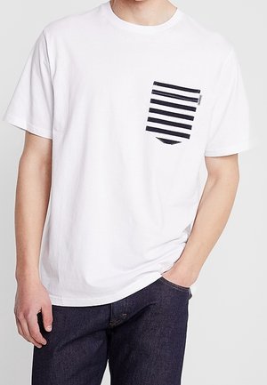 Weißes Baumwoll-T-Shirt mit kurzen Ärmeln und einer gestreiften navyblauen Tasche. Denim-Jeans ergänzen den lässigen Look, ohne zusätzliche Muster oder Akzente.