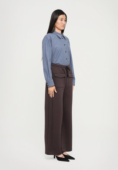 Μπλε καρό πουκάμισο με γιακά, συνδυασμένο με σκούρες καφέ παντελόνες φαρδιές με τσέπες και κορδόνι. Μαύρα γοβάκια με αιχμηρή μύτη.