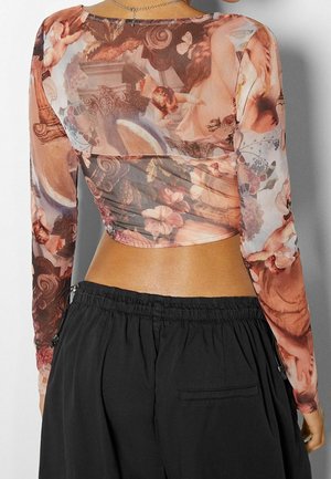 Frau trägt ein durchsichtiges, langärmeliges Crop-Top mit Renaissance-Engelchen- und Blumenmuster, kombiniert mit lockeren schwarzen Hosen.