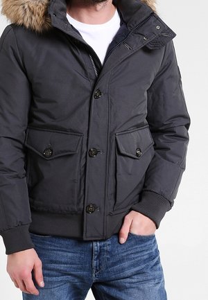 Man draagt een zwarte winterjas met bontkraag, knoop- en ritssluiting, en blauwe spijkerbroek, hand in de zak, tegen een effen achtergrond.