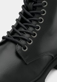 Cizmă de gleznă din piele neagră, cu ochiuri proeminente, șireturi negre și o textură netedă și lucioasă. Talpă plată cu detalii subtile de cusătură.