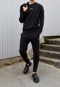 Sudadera negra con logo dorado, pantalones jogger negros a juego y zapatos slip-on negros. Tejido suave con puños y cinturilla acanalados.