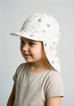 Junges Kind trägt einen weißen Sonnenhut mit pinken, gelben und grauen Herzmustern und ein beigefarbenes kurzärmeliges Hemd, blickt nach links.