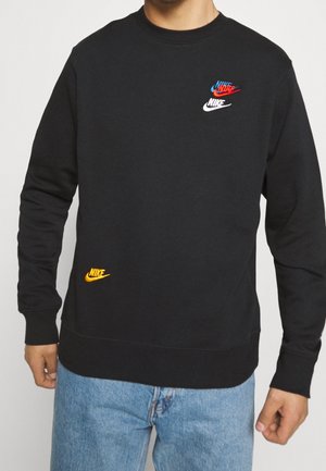 Mężczyzna w czarnej bluzie Nike z trzema małymi, wielokolorowymi logo Nike na piersi oraz żółtym logo przy talii, w połączeniu z niebieskimi dżinsami.