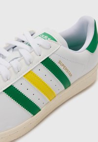 White leather sneaker featuring green and yellow zigzag-striped accents, lace-up design, and a beige textured sole. "SUPERSTAR" is printed in gold.
