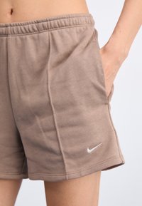 Nike šortai iš minkštos, šviesiai rudos medžiagos, su elastingu juosmeniu ir šoninėmis kišenėmis. Logotipas balta spalva yra ant apvado.