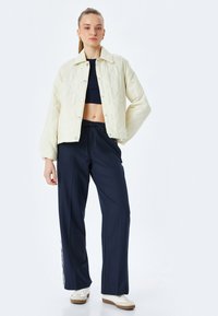 Gepolsterte hellgelbe Jacke mit Druckknopfverschlüssen, kombiniert mit marineblauen weit geschnittenen Hosen mit seitlichen Streifen und weißen Turnschuhen.