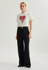 Hvid bomulds t-shirt med et rødt hjerte-design, parret med sorte højtaljede brede bukser og et sort bælte med en guldbuckle.