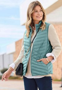 Frau, die draußen lächelt und eine türkisfarbene Steppweste, einen beigen Pullover, dunkle Hosen trägt und eine schwarze Tasche mit einer goldenen Kettenriemen in der Hand hält.