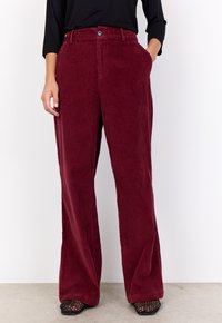 Red corduroy wide-leg pants with vertical ribbing, front pockets, and classic button closure, styled with a black top and textured shoes.