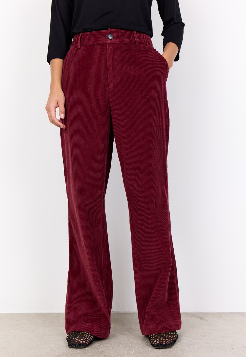 Red corduroy wide-leg pants with vertical ribbing, front pockets, and classic button closure, styled with a black top and textured shoes.