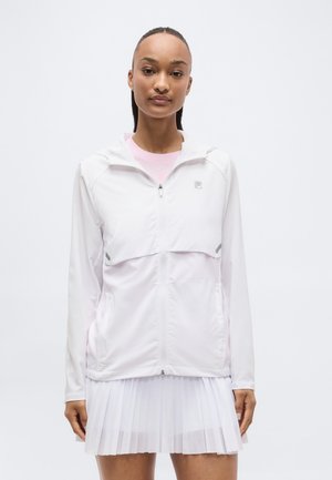 Femme portant une veste blanche zippée par-dessus un t-shirt rose et une jupe plissée blanche, debout devant un fond clair uni.