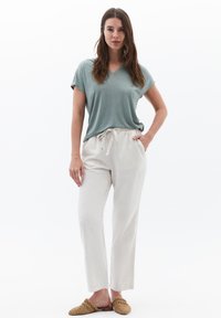 Camiseta de rayón verde claro con cuello en V y mangas cortas, combinada con pantalones crema con cordón y zapatos tipo mocasín de tejido beige. Texturas suaves.