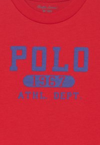 Červené bavlněné tričko s modrým blokovým textem: "POLO 1967 ATHL. DEPT." Kolečkový designový prvek za textem "1967".