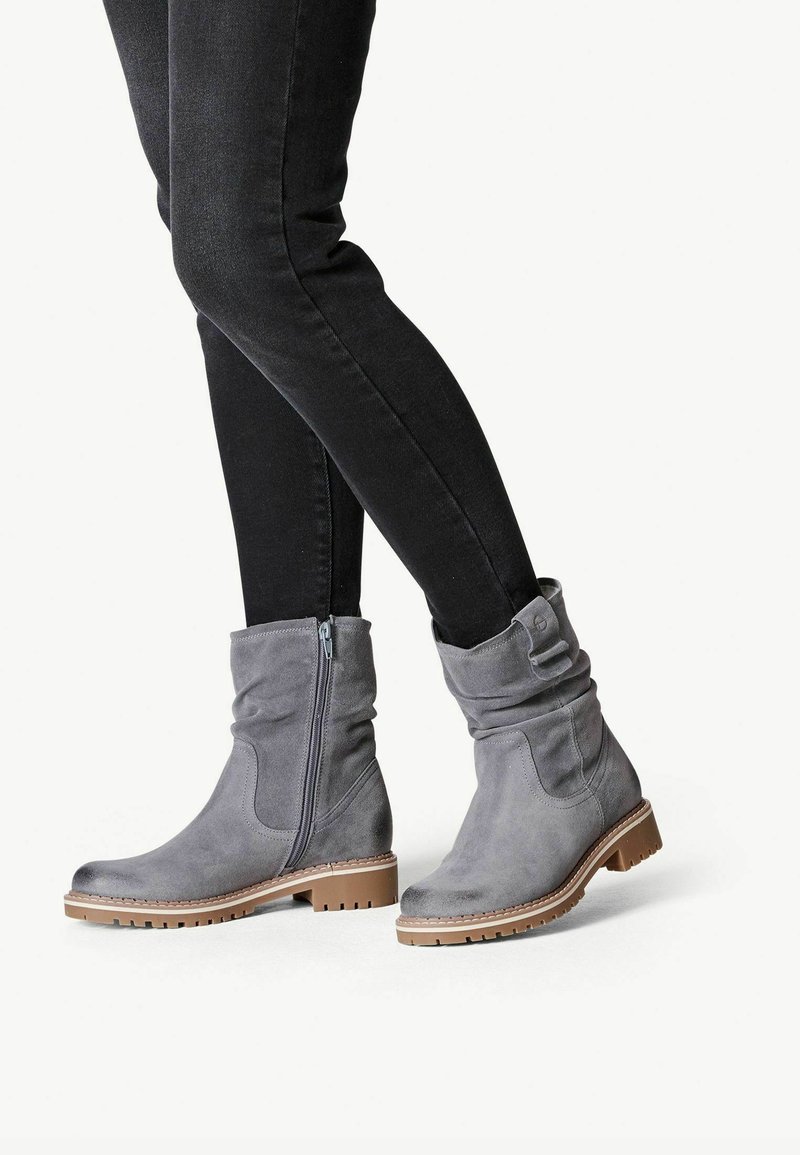 Personne portant un pantalon noir ajusté et des bottines en daim gris avec fermeture éclair latérale et semelles crantées basses marron, sur fond blanc.