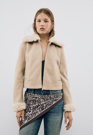 Jeune femme portant une veste beige à col en fourrure, un jean bleu et une écharpe à imprimé zèbre nouée autour de la taille, sur un fond uni.