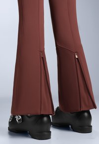 Hnedé rozšírené lyžiarske nohavice majú zipsy na spodnej časti nôh a sú skombinované s čiernymi lyžiarskymi topánkami s výrazne textúrovaným povrchom.