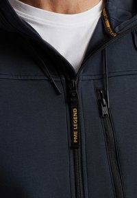 Marineblå zip-up hoodie lavet af blødt stof, med en høj krave, gult logo på lynlåsen og en struktureret overflade med kontrasterende detaljer.