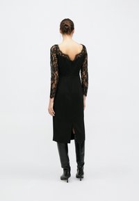 Robe noire avec manches longues en dentelle, coupe ajustée, dos en V profond, longueur genou et fente latérale. Le modèle porte des bottes noires montantes jusqu'au genou.