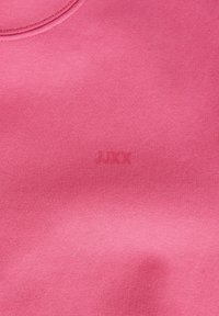 Gros plan sur un tissu rose montrant une texture fine en maille et un logo "JUXX" subtilement embossé près de la couture arrondie du col.