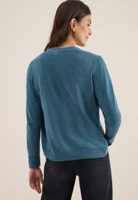 Pull en maille teal avec un col rond, des manches longues et un ourlet côtelé, associé à un jean sombre. Texture lisse, coupe décontractée et design minimaliste.