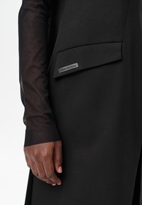 Donkere hand met een doorschijnende zwarte mesh mouw naast een zwart kledingstuk met een label "blauwe schaduw" op een klepzak.