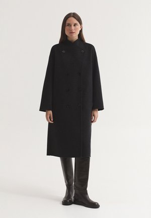 Femme debout portant un long manteau noir croisé et des bottes hautes sombres sur un fond clair uni.