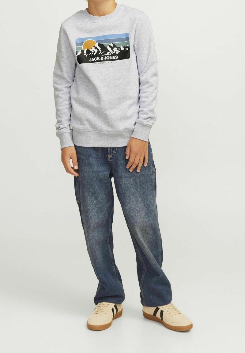 Světle šedá mikina s grafickým designem zobrazujícím hory a slunce; v kombinaci s tmavě modrými volnými džíny a béžovými teniskami s černými pruhy.