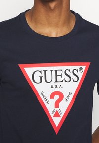 T-shirt in cotone blu navy con una grafica triangolare rossa e bianca. Presenta il logo "GUESS U.S.A.", le scritte "WASHED" e "JEANS" e un dettaglio a forma di punto interrogativo.