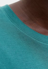 Gros plan sur un cou à peau foncée portant un t-shirt à col rond bleu sarcelle, montrant la texture du tissu et les détails de la couture.