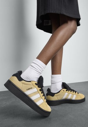 Person med gula Adidas Campus-sneakers med svarta sulor, vita ränder, svarta snören och vita strumpor, stående på grått golv.