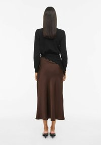 Femme aux cheveux raides et foncés portant un haut noir à manches longues, une jupe midi marron avec bordure en dentelle noire, et des chaussures à talons chaton noires, debout.