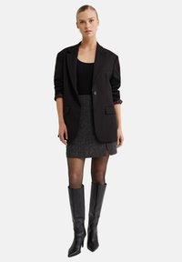 Blazer noir ajusté sur un haut noir cintré, mini-jupe en tweed gris avec fente sur le côté, et bottes en cuir noires à talons aiguilles jusqu'aux genoux.
