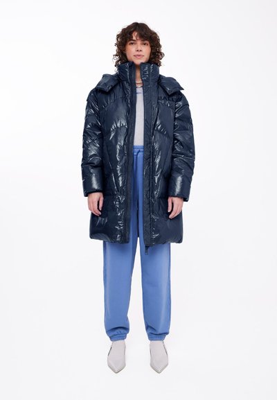 Mornarsko modra puhovka z bleščečo obdelavo, visokim ovratnikom in zadrgo. V kombinaciji s svetlo modrimi hlačami in ostrimi svetlo sivimi čevlji.