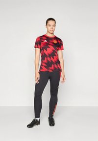 Camisola vermelha e preta com padrão em ziguezague, com mangas curtas e decote redondo. Leggings pretas com riscas vermelhas laterais e chuteiras de futebol.