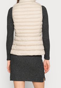 Beige gesteppte Weste mit hohem Kragen, leichtes Material, kombiniert mit einem dunkelgrauen Rippkleid, das ein enganliegendes, strukturiertes Design zeigt.
