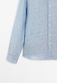 Light blue linen shirt with long sleeves, button closure, and a slightly curved hem. Soft texture with a subtle woven pattern.