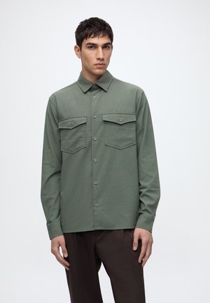 Jeune homme portant une chemise kaki à manches longues boutonnée avec poches poitrine, debout devant un fond clair uni.